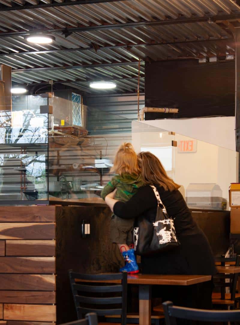 Woman holding a young child while looking at the counter inside Bagel Chicks, showing the family-friendly atmosphere.