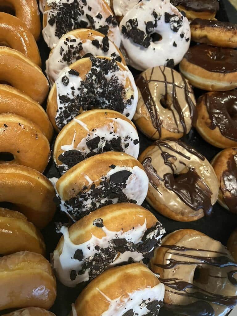 Assorted frosted donuts and chocolate-glazed treats in a tray, highlighting Bagel Chicks growing and expanding to a new location for the community.