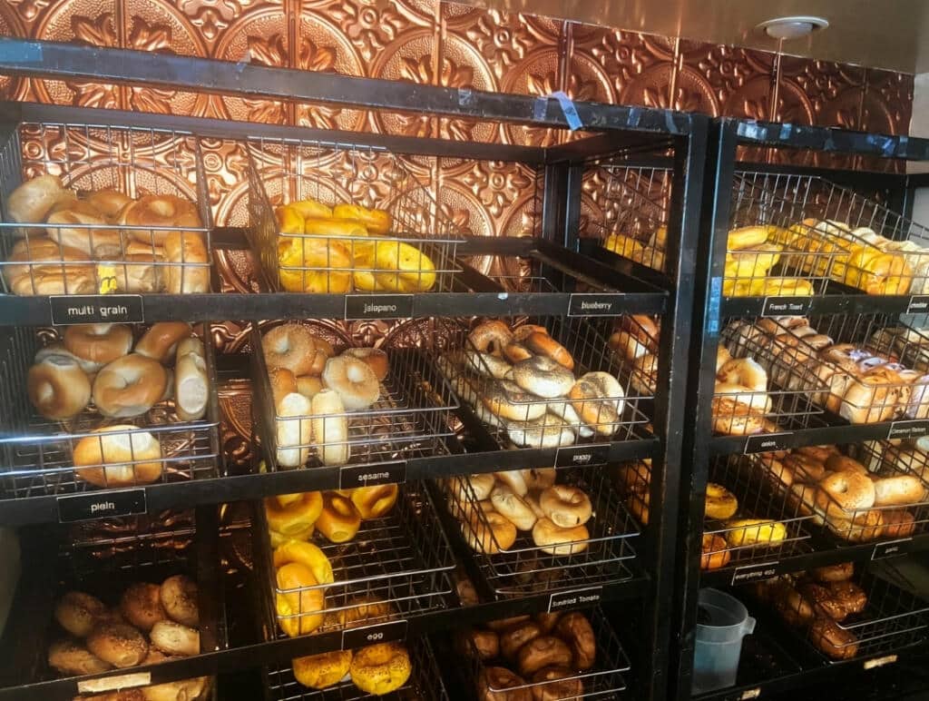 Wall of bagel racks labeled multigrain, jalapeño, blueberry, French toast, sesame, poppy, onion, plain, egg, and sundried tomato—illustrating the real history of the bagel and why it still matters to us.