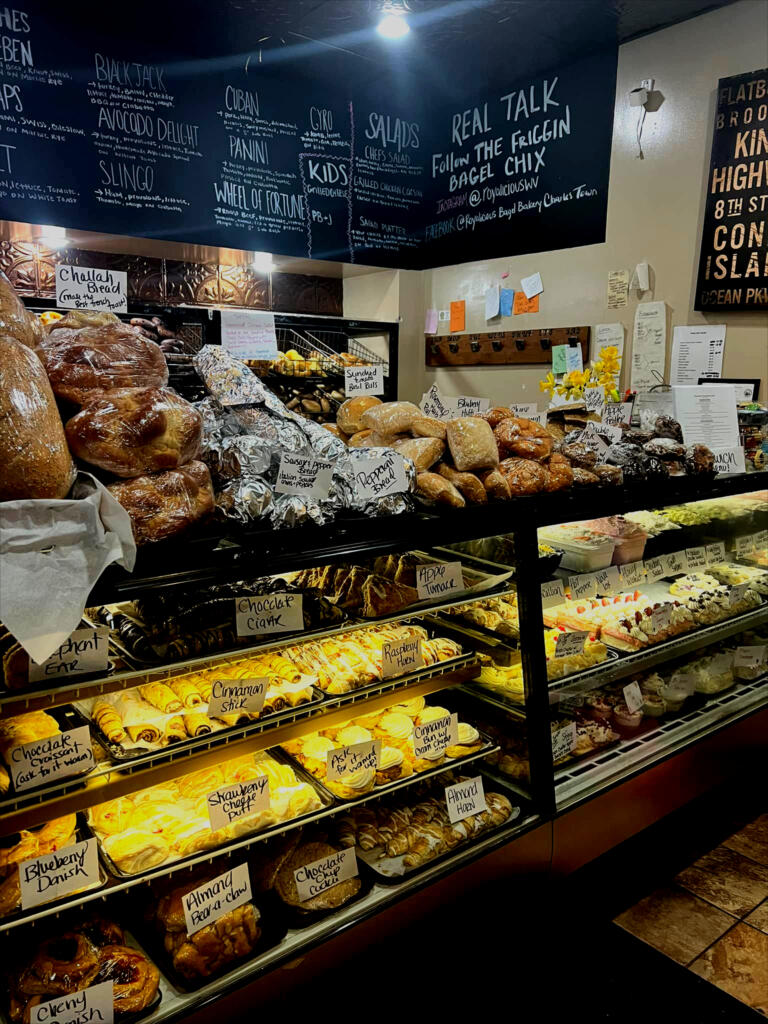 Wide view of the Bagel Chicks bakery counter with breads, pastries, desserts, and menu boards in a warm breakfast shop setting.