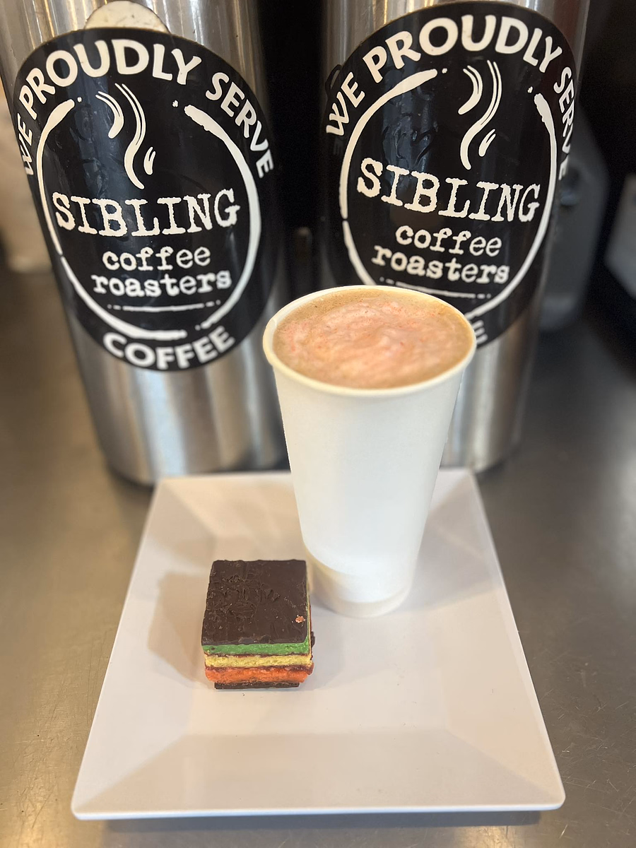 A hot coffee in a white cup served with a small layered chocolate dessert on a white plate, with Sibling Coffee Roasters containers in the background.
