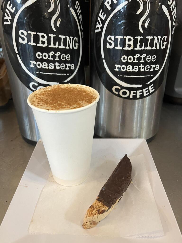 Hot coffee and a biscotti in front of Sibling Coffee Roasters containers, representing what to bring to your morning office meeting that people will actually eat.