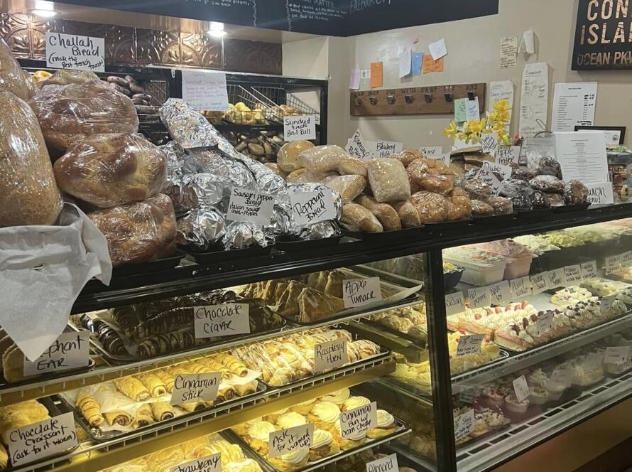 A Bagel Chicks bakery display filled with bagels, breads, pastries, danishes, cakes, and deli items under a chalkboard menu, perfect for planning a spring picnic order.