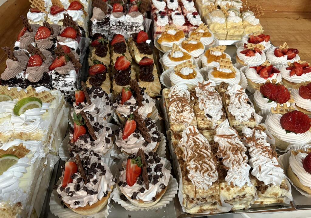 Assorted mini cakes, cream pastries, strawberry tarts, chocolate desserts, and fruit-topped sweets arranged in a bakery display, ideal for a morning office meeting.